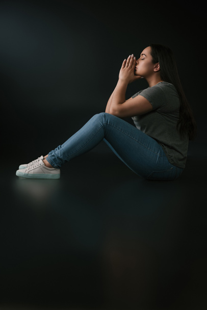 Side view of upset plus size girl covering face with hands and sitting on black background - Photo, Image
