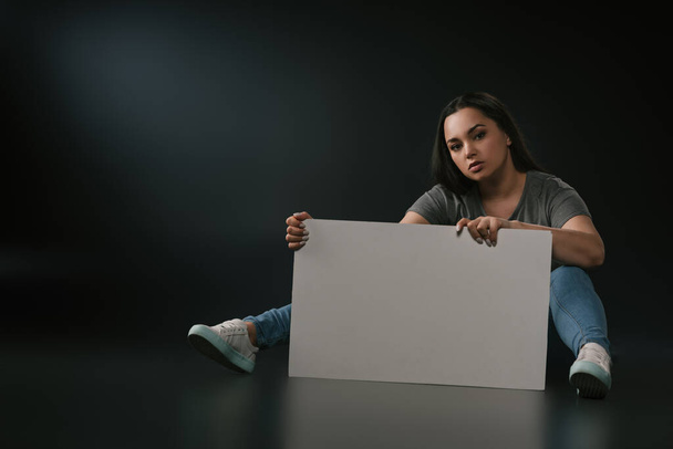 Plus size girl sitting with blank placard on black background - Foto, imagen