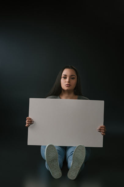Front view of beautiful plus size girl sitting with empty board on black background - Foto, immagini