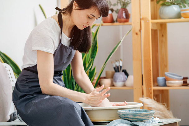 The concept of home hobbies. Woman in apron prepares to work behind a potter's wheel, sets a clay ball on it - 写真・画像