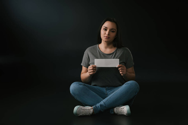 Front view of peaceful plus size girl holding empty card and sitting on black background - Photo, Image