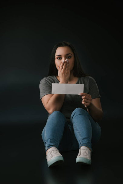 Front view of plus size girl covering mouth with hand and holding empty card on black background - Fotoğraf, Görsel