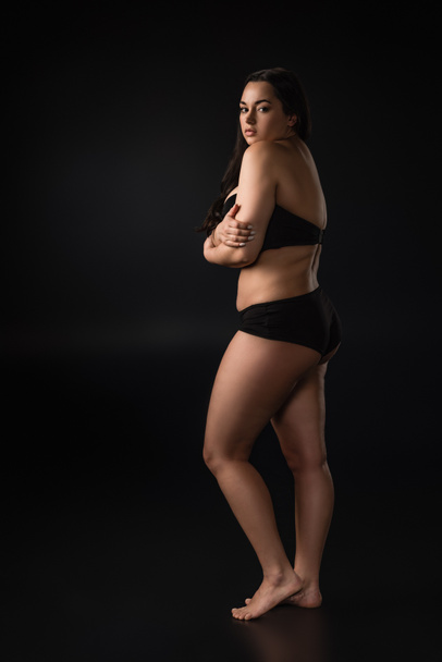 Вид в полный рост девушки plus size, скрывающей тело со стыдом на черном фоне
 - Фото, изображение