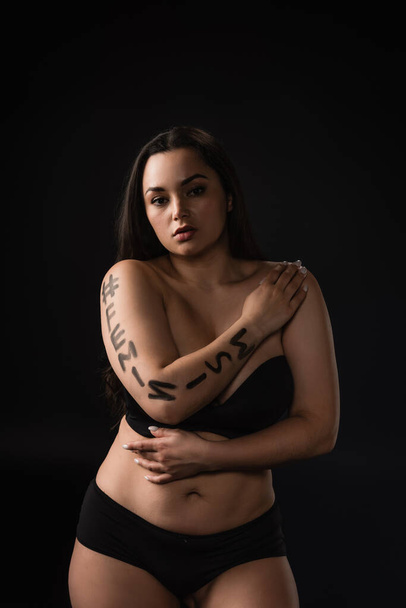 Front view of plus size model in black underware with lettering hashtag feminism on body isolated on black - Foto, immagini