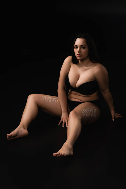 Plus size model in underwear with lettering I Did not Ask For It on body sitting isolated on black  - Photo, Image