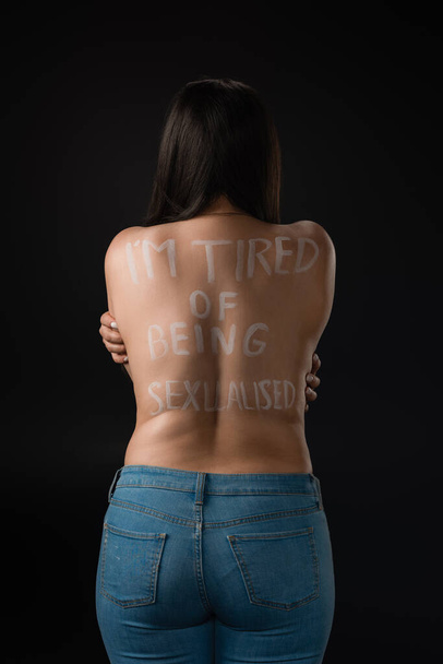 Back view of plus size model with lettering I Am Tired of Being Sexualised on naked back isolated on black  - Fotó, kép