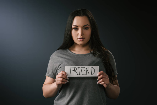 Вид спереди красивой plus size девушки, держащей открытку со словом friend на чёрном фоне
 - Фото, изображение