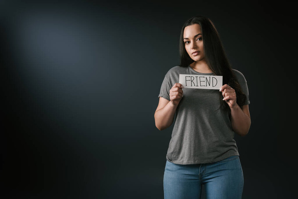 Front view of brunette plus size girl holding card with word friend on black background - Fotografie, Obrázek