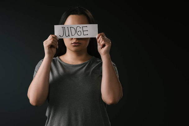 Front view of plus size girl covering eyes with card with word judge on black background - Photo, Image