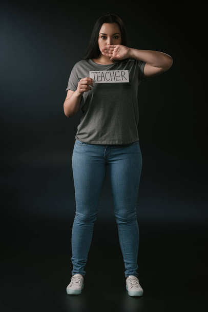 Full length view of plus size κορίτσι κρατώντας κάρτα με τη λέξη δάσκαλος και καλύπτοντας το στόμα με το χέρι σε μαύρο φόντο - Φωτογραφία, εικόνα