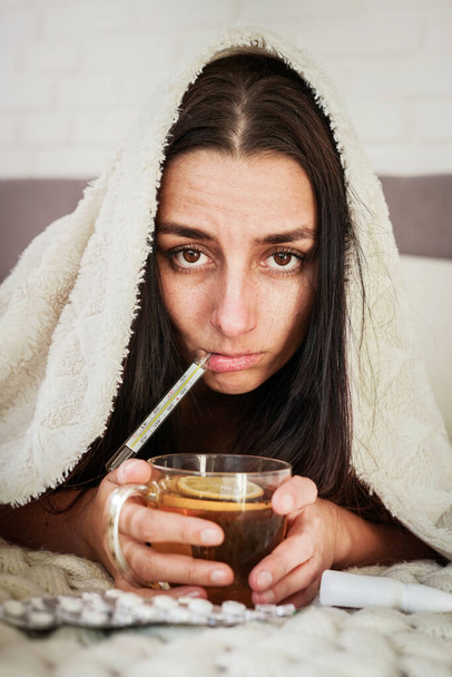 Young woman is feeling bad and sick lying in the bed at home. Girl Has temperature and drink tea with pillsGirl has symptoms of respiratory viral infection, fever, cough, headache shortness of breath. Coronavirus epidemic pandemic concept. - Foto, Imagem