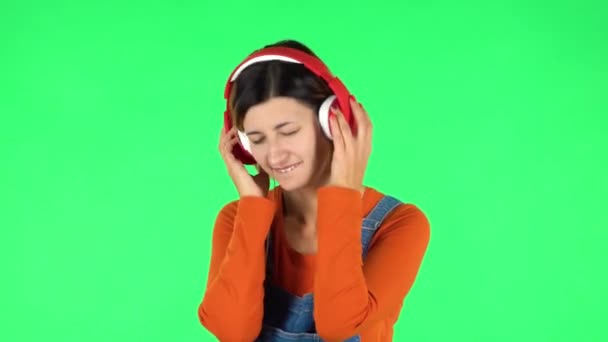 Cheerful girl dancing and enjoys music in big red headphones. Green screen - Πλάνα, βίντεο