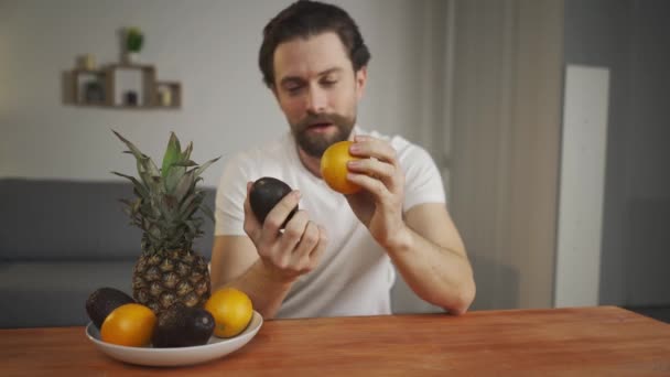 Ein junger Mann sitzt an einem Tisch und spricht über die Nützlichkeit von Früchten, er hebt sie auf und zeigt Ananas, Avocado, Orange. Fokus auf Früchte - Filmmaterial, Video