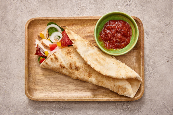 top view of fresh burrito with chicken and vegetables on board near chili sauce on concrete grey background - Photo, Image