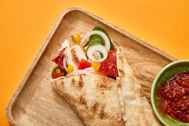 top view of fresh burrito with chicken and vegetables on board near chili sauce on orange background - Fotoğraf, Görsel