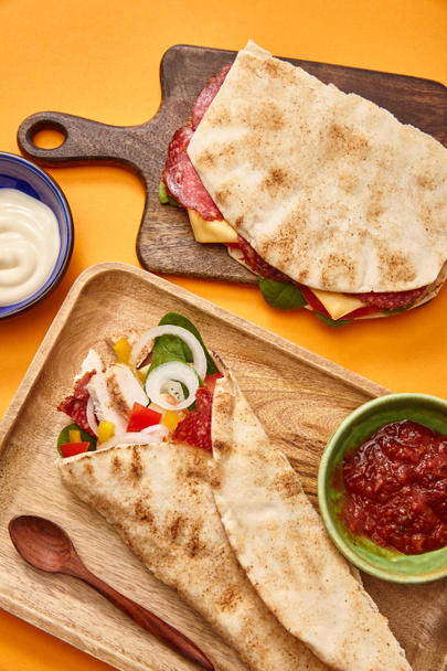 τοπ άποψη του φρέσκου σάντουιτς με σαλάμι σε πίτα και burrito με κοτόπουλο σε σανίδες κοντά σάλτσες σε πορτοκαλί φόντο - Φωτογραφία, εικόνα