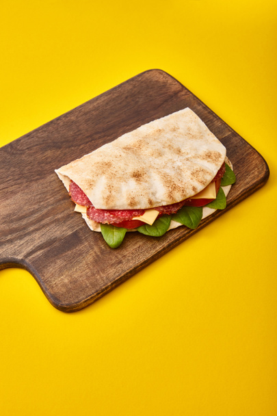 čerstvý sendvič se salámem, pitou, zeleninou a sýrem na dřevěné řezací desce na žlutém pozadí - Fotografie, Obrázek