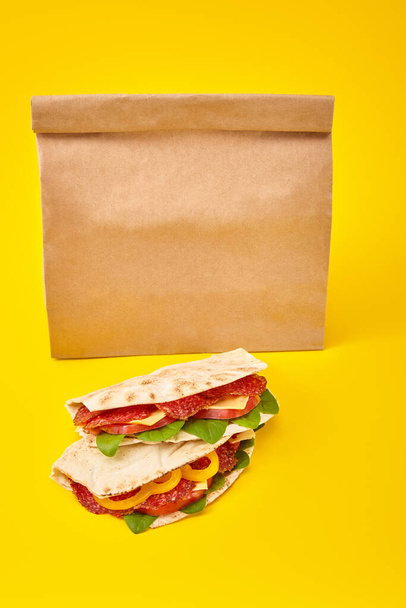 fresh sandwiches with salami, pita, vegetables and cheese on yellow background near paper takeaway bag - Fotografie, Obrázek