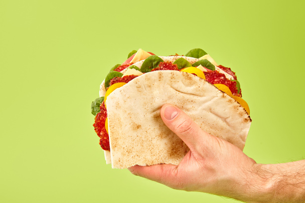 partial view of man holding fresh sandwich with salami, pita, vegetables and cheese isolated on green - Photo, Image