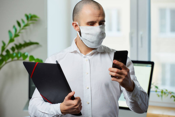 A man wearing a protective mask against a virus. Doctor with a surgical mask against the 2019-nCoV using the phone and the other hand holding a folder with files in his office. Coronavirus quarantine. - Фото, изображение