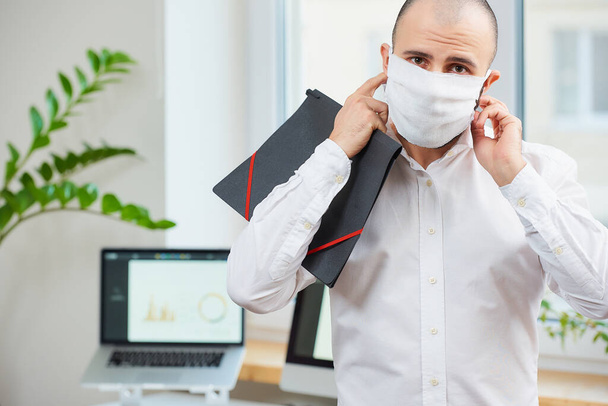 Ein Mann, der eine Gesichtsmaske gegen das Coronavirus abnimmt. Ein Büroangestellter hält an seinem Arbeitsplatz einen Ordner mit Computern und grünen Pflanzen im Hintergrund. Die COVID-19-Quarantäne. - Foto, Bild
