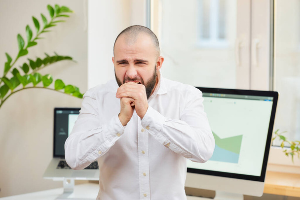 Egy fehér inges, szakállas férfi erősen köhög az öklébe. Egy irodai munkás a munkahelyén, számítógépekkel és zöld növényekkel a háttérben. Koronavírus (COVID-19) karantén. - Fotó, kép
