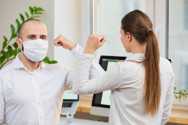 Ellenbogengruß zur Vermeidung der Ausbreitung des Coronavirus (COVID-19). Ein Mann mit medizinischer Gesichtsmaske und ein Mädchen treffen sich mit bloßen Händen in einem Büro. Statt mit einer Umarmung oder einem Händedruck begrüßen sie sich mit den Ellbogen - Foto, Bild