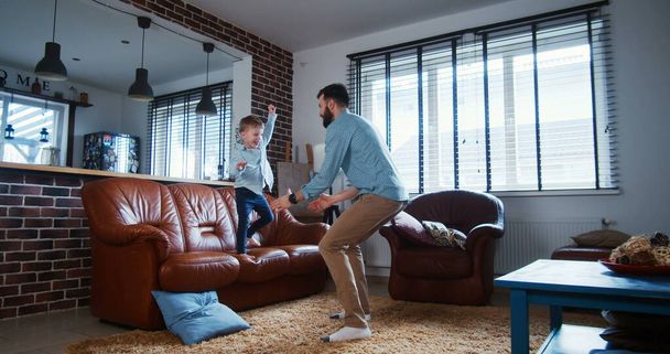 Happy young Caucasian father lifting his little son high, playing and having fun in modern light living room slow motion - Zdjęcie, obraz