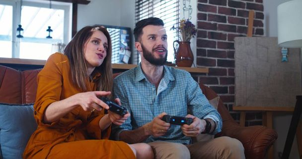 Νέοι χαρούμενοι Καυκάσιοι σύζυγοι παίζουν βιντεοπαιχνίδια ο ένας εναντίον του άλλου στο σπίτι απολαμβάνοντας τον ελεύθερο χρόνο τους σε αργή κίνηση. - Φωτογραφία, εικόνα