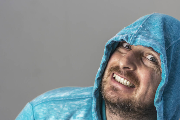 Portrait of a cunning man with unshaven face dressed in blue hoodie. Gray background. Close-up. - Photo, Image