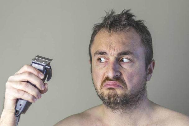Porträt eines haarigen Mannes mit unrasiertem Gesicht, der einen elektrischen Haarschneider hält und ihn misstrauisch betrachtet. - Foto, Bild