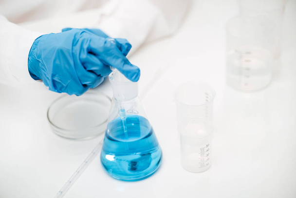 Лаборант в гумових рукавичках, що вказує на колбу з синьою рідиною на столі
 - Фото, зображення