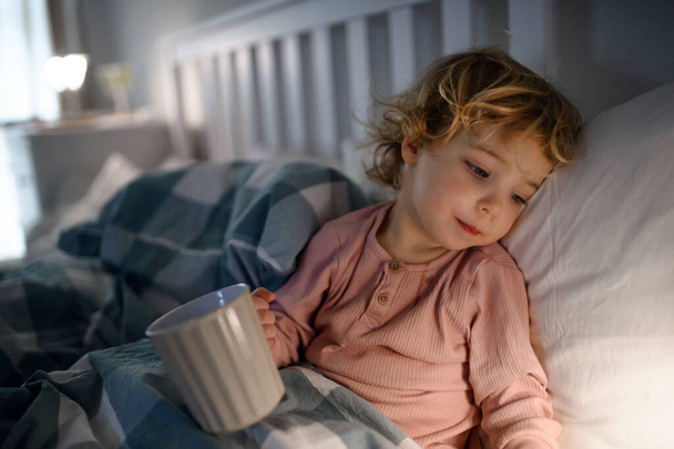 Маленькая больная девочка лежит в постели дома, держа чашку
. - Фото, изображение