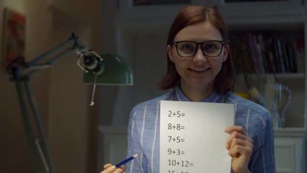 30s woman in glasses showing sheet of paper with simple mathematical equations looking at camera at home. Online education process. Webcam view of teacher talking.  - Metraje, vídeo