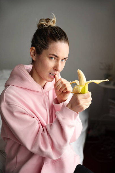 Young woman sitting in bed, eating banana and resting. Vertical picture. - Фото, зображення