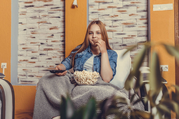Uzaktan kumandayla TV seyreden ve patlamış mısır yiyen rahat bir genç kız evdeki rahat koltukta oturuyor.. - Fotoğraf, Görsel