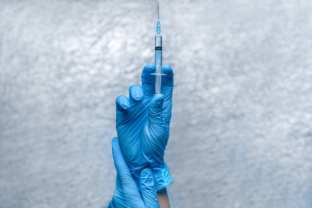 Mavi koruyucu eldivenli eller, üzerinde koronavirüs virüsü bulunan bir şırınga tutuyor. Arka planda gri bir aşı var.. - Fotoğraf, Görsel