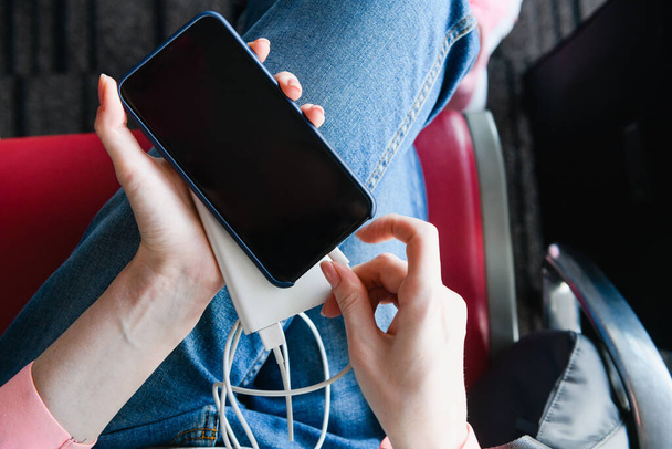 Женщина в джинсах на красном терминале аэропорта держит портативное зарядное устройство, подключенное к мобильному телефону, чтобы пополнить аккумулятор, до просмотра черного заблокированного экрана. Вид сверху
. - Фото, изображение