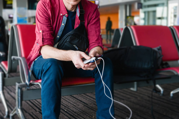 Man tourist hold smartphone charging by usb cable from portable power bank, sitting with luggage at airport gate - Photo, Image