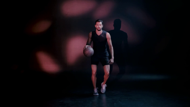 Handsome sportsman playing basketball on dark background - Footage, Video