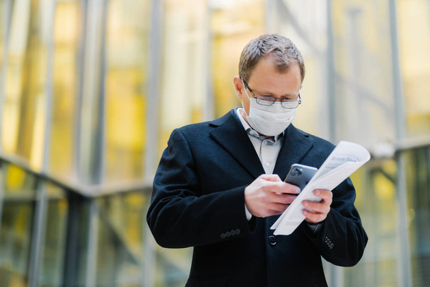 Coronavirus pandemic concept. Photo of male manager focused in smartphone device, sends text messages, wears spectacles and medical mask, holds papers, walks at street during quarantine, pandemic - Foto, Imagen