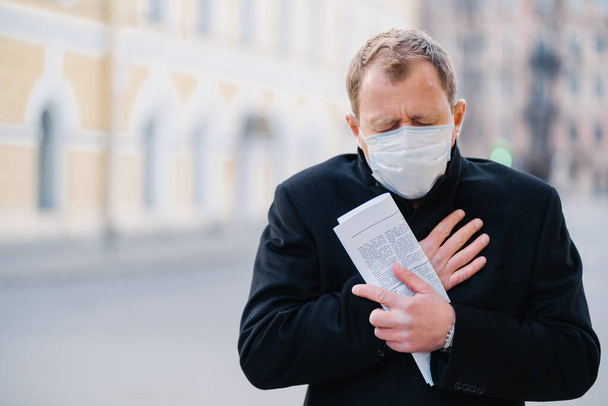 Zarażony człowiek nosi maskę medyczną, ma ciągły kaszel, objawy Covid-19, trzyma zwiniętą gazetę, pozuje na mieście na świeżym powietrzu, potrzebuje izolacji, aby zatrzymać rozprzestrzenianie się Coronavirus. Środki zapobiegawcze - Zdjęcie, obraz