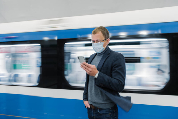 Photo of man in eyewear and protective medical mask, uses modern phone, travels by public transport, stands against train in motion on platform, gets information online about new Covid-19 virus - Fotó, kép