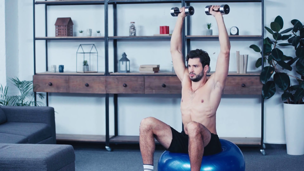 young shirtless sportsman sitting on fitness ball and training with dumbbells - Imágenes, Vídeo