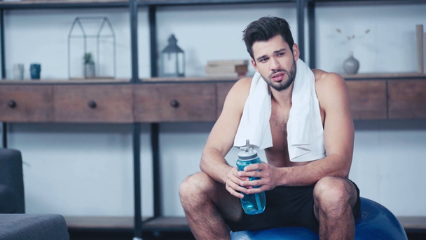 desportista cansado com toalha no pescoço sentado na bola de fitness e água potável
 - Filmagem, Vídeo