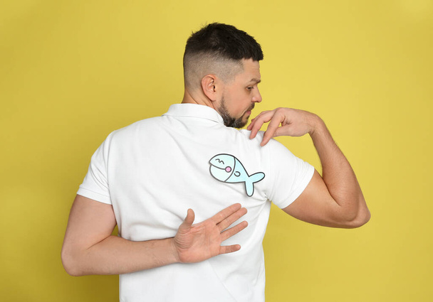 Man with paper fish on back against yellow background. April fool's day - Φωτογραφία, εικόνα