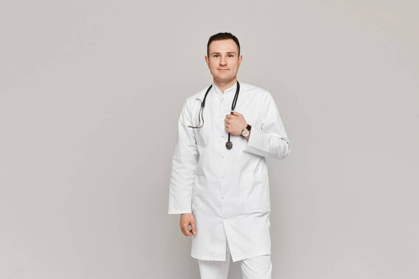 Young male doctor in medical uniform posing with stethoscope on grey background. Concept of healthcare - Zdjęcie, obraz