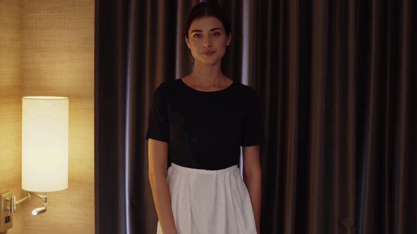 beautiful, young maid in white apron smiling at camera in hotel room - Video