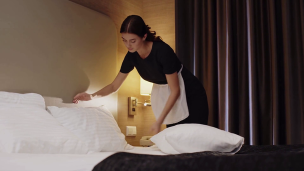 joven, hermosa criada en uniforme y delantal haciendo cama en habitación de hotel
 - Imágenes, Vídeo