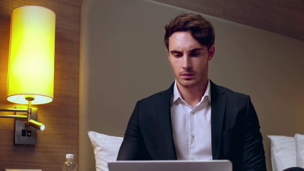 przemyślany, młody biznesmen korzystający z laptopa w pokoju hotelowym - Materiał filmowy, wideo